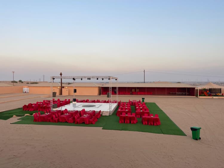 Outdoor desert event setup with empty stage, surrounded by red chairs and tables on green artificial turf. 