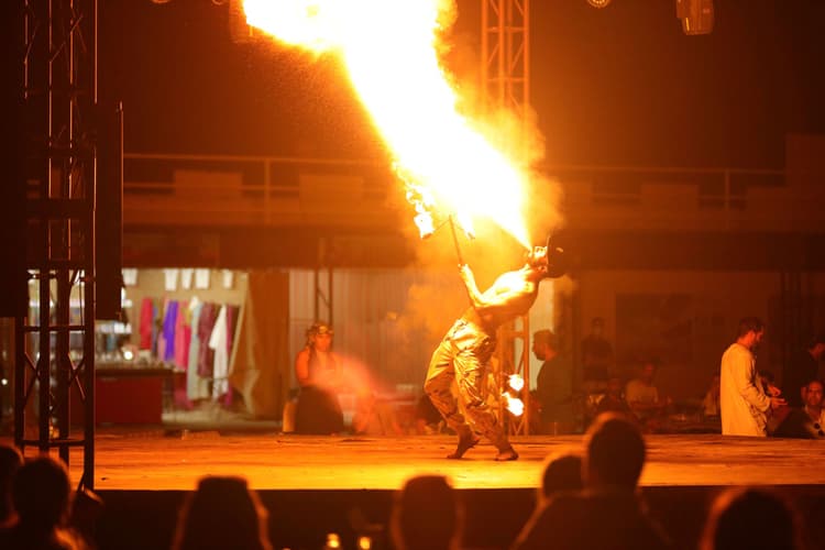 A fire performer breathes flames on a dimly lit stage, surrounded by an attentive audience.