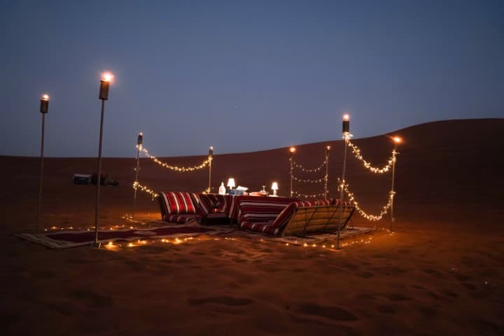 A cozy desert picnic at dusk with striped cushions and a small table. String lights and torches