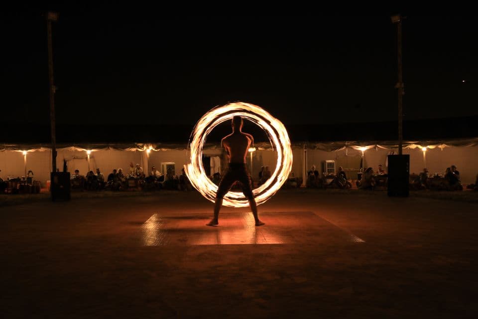 A fire performer spins a blazing circle at night, surrounded by a dimly-lit audience.