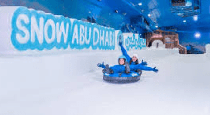 Two people joyfully ride an inflatable tube down a snowy indoor slope at Snow Abu Dhabi, with blue toned walls and large lettering in the background.