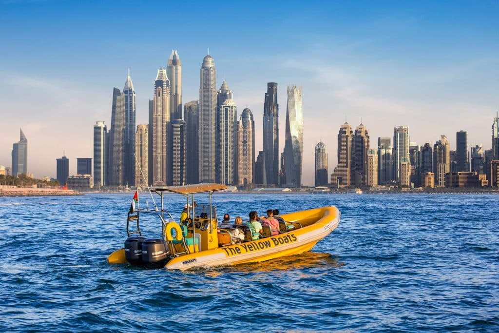 A yellow tour boat with people on board sails in calm blue water, set against a backdrop of a modern city skyline
