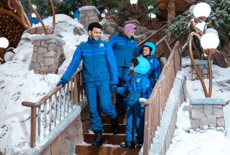 Snow Abu Dhabi: A family in blue winter jackets and helmets happily descends snowy stone steps, surrounded by winter decorations and glowing lanterns