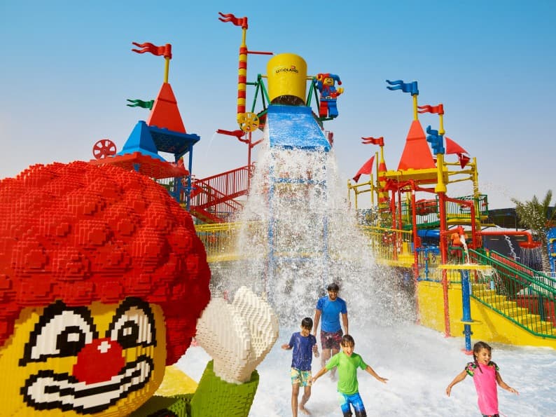Children enjoying a water park with colorful slides and a large tipping bucket. A giant Lego figure waves in the foreground.