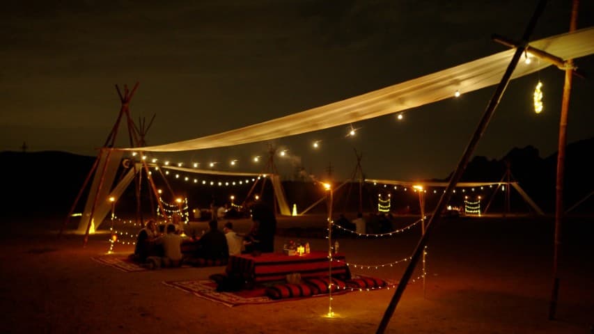A tent adorned with lights and candles, set against a serene desert backdrop, creating a cozy dining atmosphere.