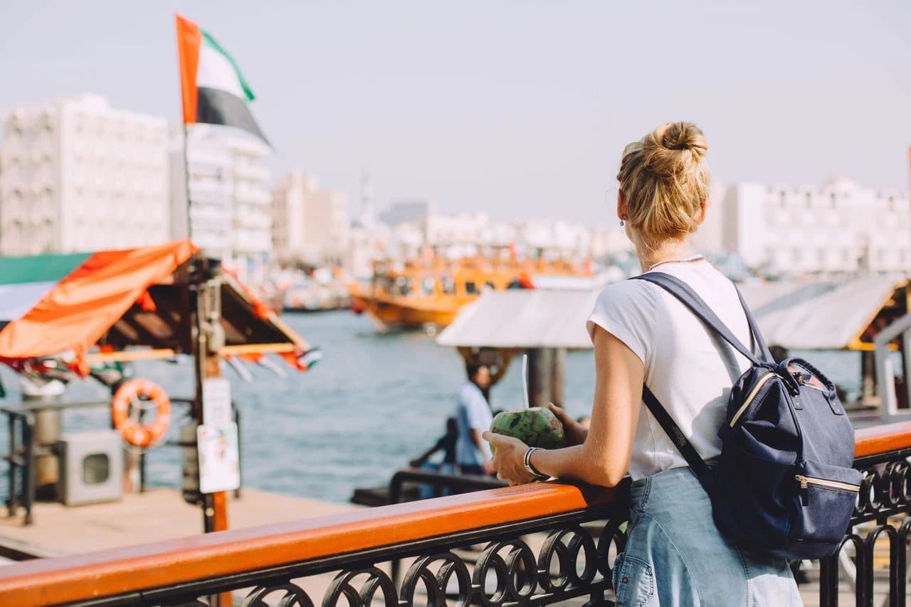 A woman with a backpack gazes at boats on a sunny waterfront. She holds a coconut