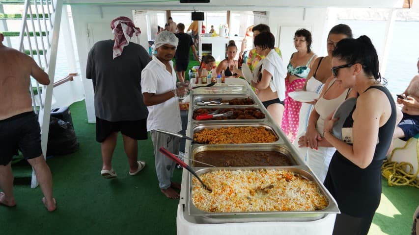People in swimwear enjoy a buffet on a boat, featuring colorful dishes like rice and curries.