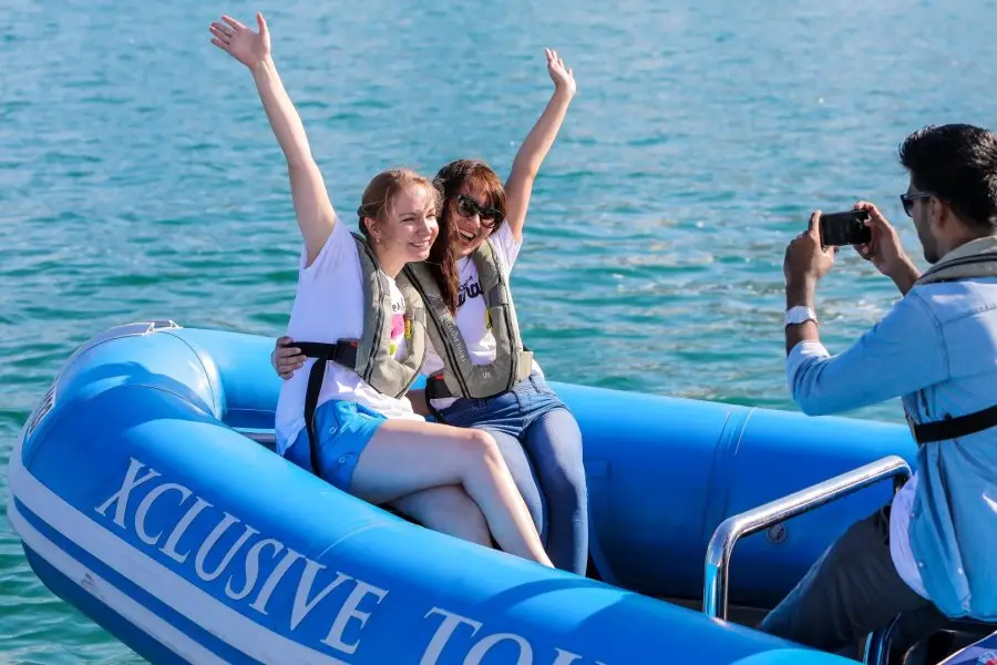 Two women joyfully raise their arms on a blue inflatable boat labeled "Xclusive Tours." 