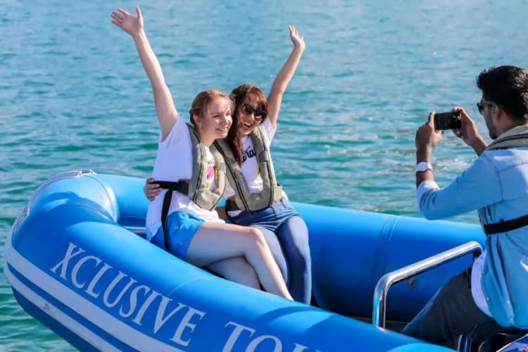 Two women joyfully raise their arms on a blue inflatable boat labeled "Xclusive Tours." 