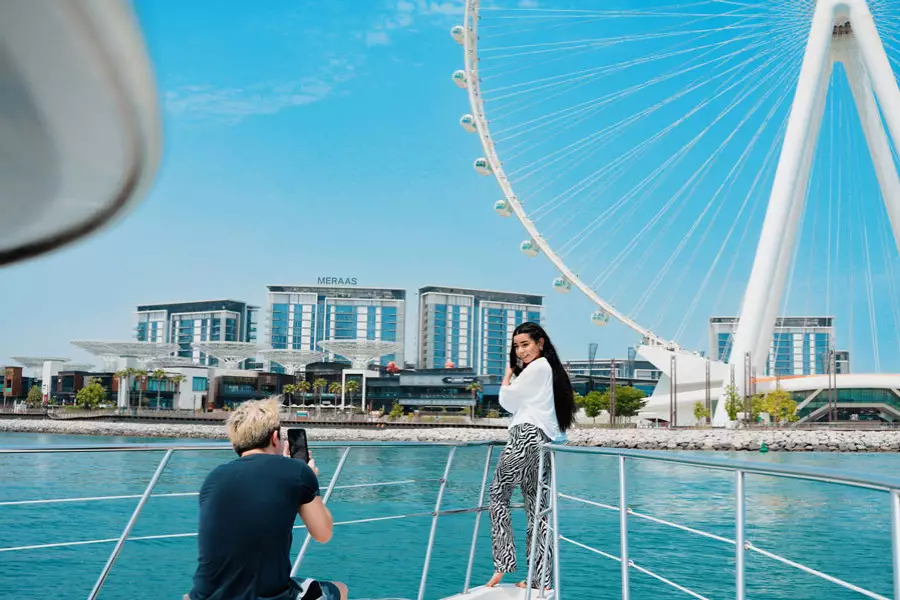 A woman poses on a boat against a blue sky, with a large Ferris wheel and modern buildings in the background. 