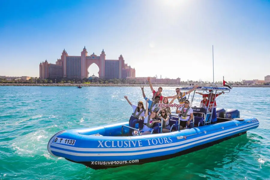 A group of people on a blue "Xclusive Tours" speedboat cheerfully wave, with a large, ornate hotel in the background. 