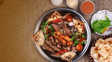 A platter of mixed grilled meats with vegetables and flatbreads, surrounded by small bowls of sauces and fresh greens.