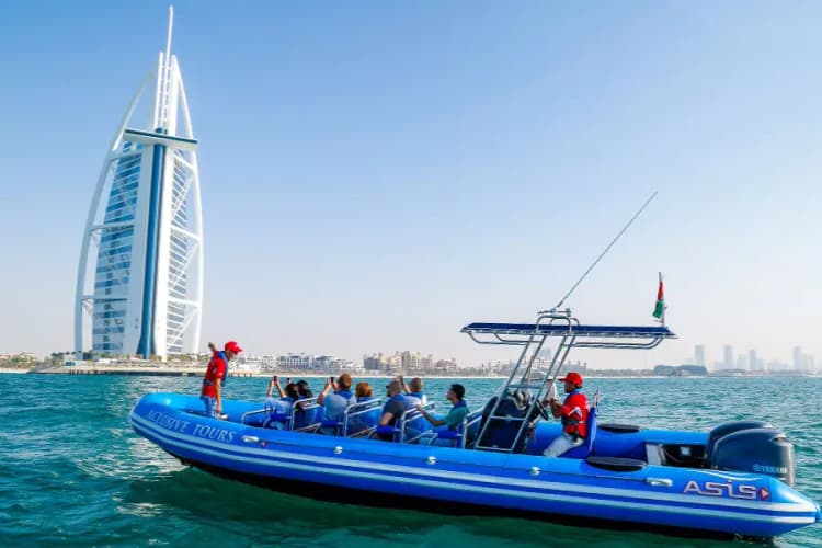 A blue tour boat with passengers cruises on calm waters. The iconic sail-shaped Burj Al Arab hotel stands prominently in the background 