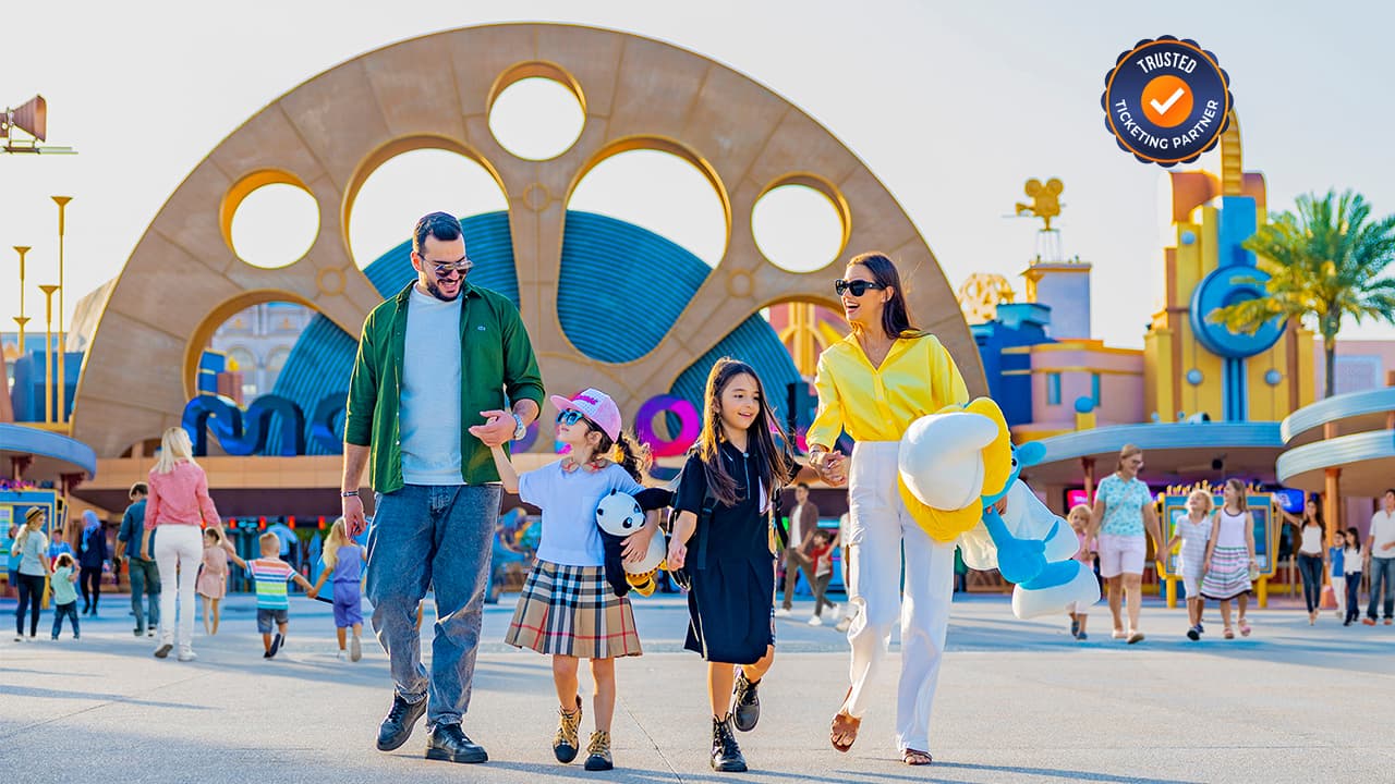 A smiling family of four joyfully walks hand-in-hand at Motiongate park entrance. The backdrop features large, colorful, circular designs.