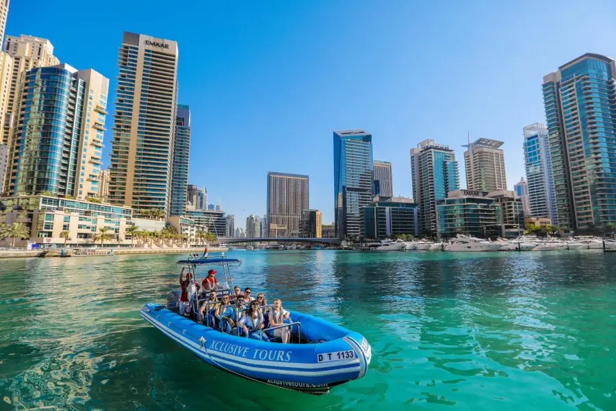 A blue tour boat with smiling passengers floats on turquoise water, framed by modern skyscrapers under a clear blue sky