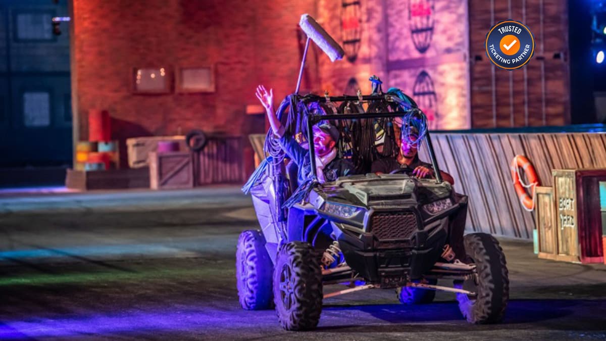 Two people in colorful costumes drive a rugged ATV on a vibrantly lit stage with a playful, adventurous atmosphere. 