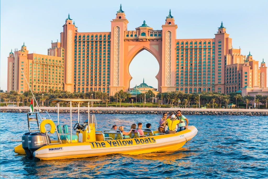A yellow tour boat on blue water with passengers enjoying views of a large, lavish resort.