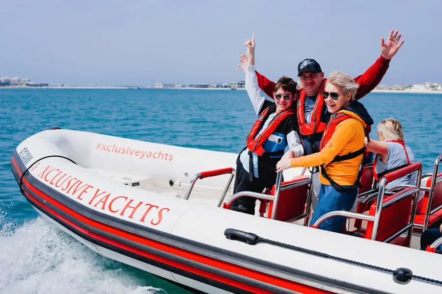 Three people smile and wave on a speeding "Xclusive Yachts" boat in blue waters