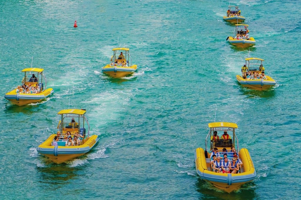 Nine yellow speedboats with tourists navigate turquoise waters in a line, creating small waves.