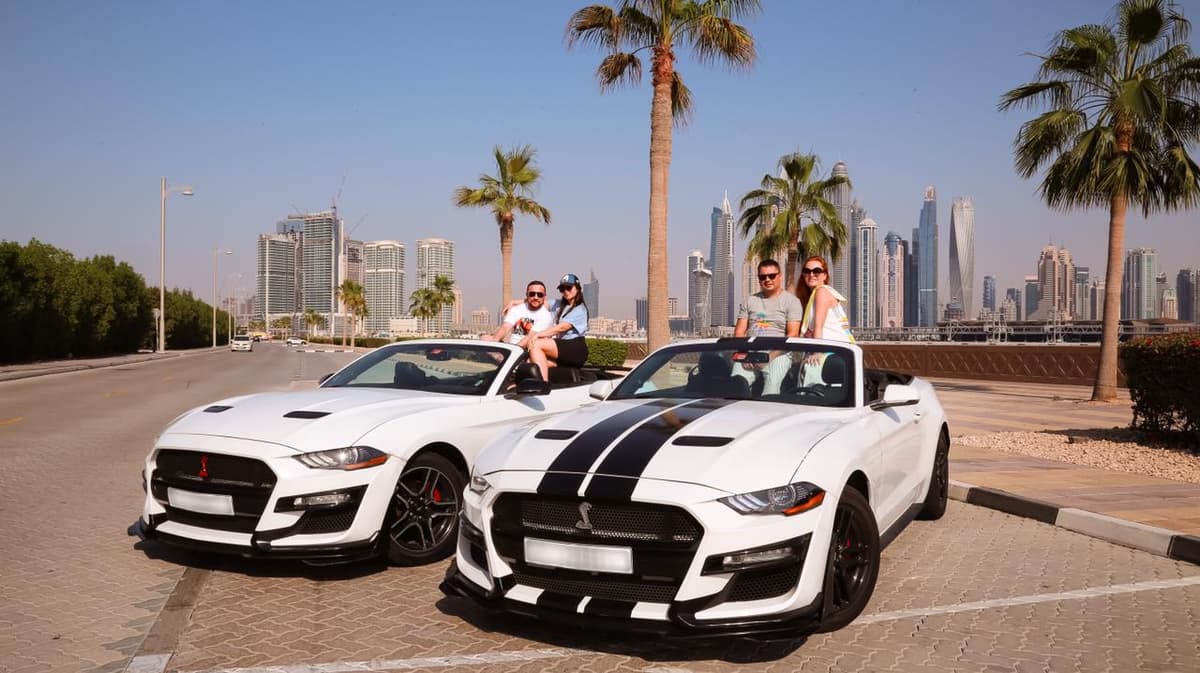 Two white sports cars with black stripes are parked on a sunny street. Four people are joyfully posing with the cars.