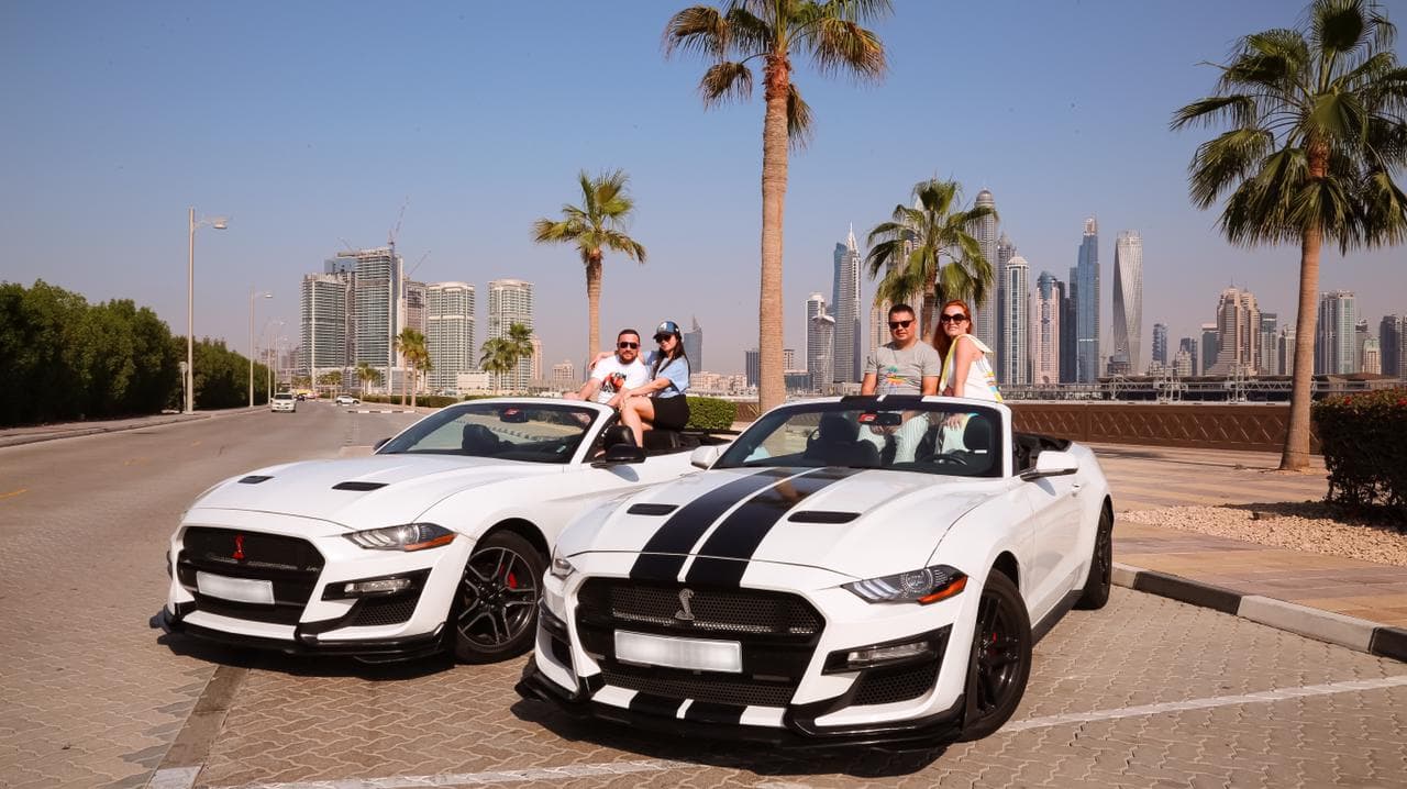 Two white sports cars with black stripes are parked on a sunny street. Four people are joyfully posing with the cars.