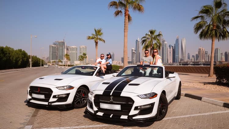 Two white sports cars with black stripes are parked on a sunny street. Four people are joyfully posing with the cars.