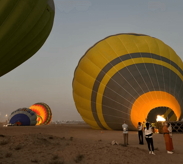 Hot air balloons being inflated on a desert at dawn. People stand nearby