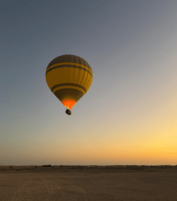 A yellow hot air balloon with dark stripes floats gently in a clear sunset sky over a vast, barren desert landscape,