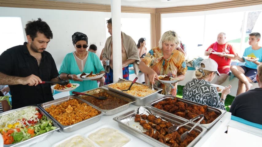People serving themselves at a buffet with various dishes, including salads and grilled meats, on a boat. 