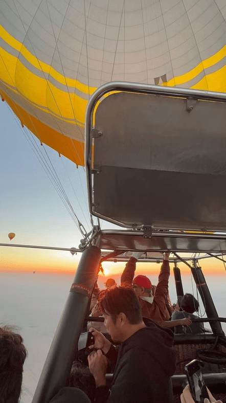 People in a hot air balloon basket admire a vibrant sunrise, with one person taking a photo. 
