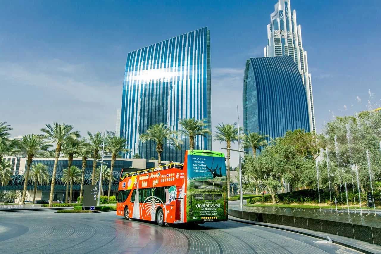 A vibrant red tour bus drives past modern skyscrapers and palm trees under a clear blue sky.