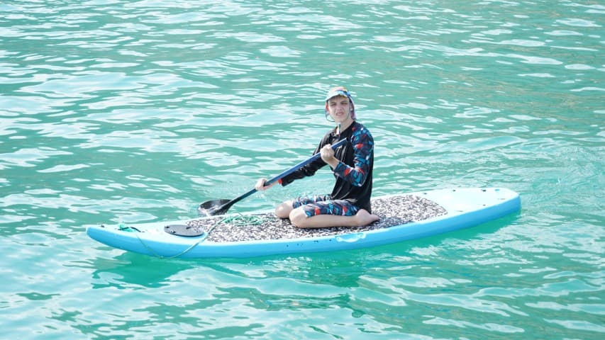 A person is sitting on a blue paddleboard in clear turquoise water, smiling and holding a paddle