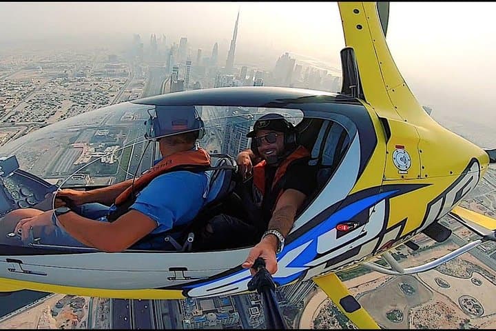 Two people take a selfie in a gyrocopter, flying over a cityscape with buildings and streets visible below.