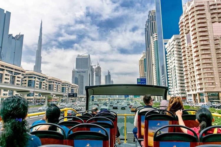 People sit on the upper deck of an open-top bus touring a modern cityscape with skyscrapers under a partly cloudy sky