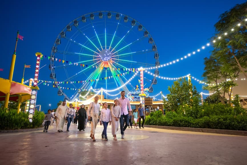 People walks at an amusement park at dusk. A brightly lit Ferris wheel and festive string lights create a joyful, vibrant atmosphere.