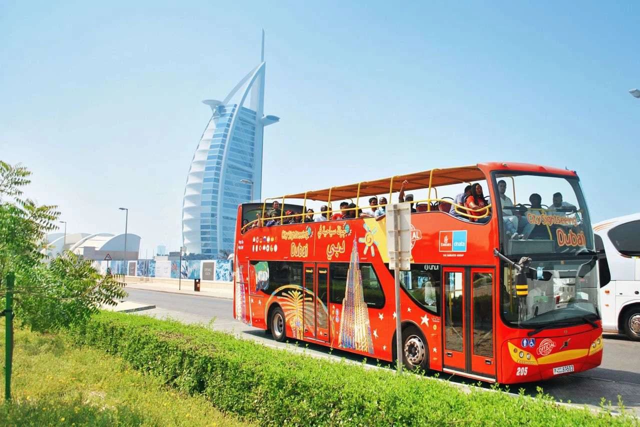 A bright red double-decker tourist bus in Dubai, with the iconic sail-shaped skyscraper in the background