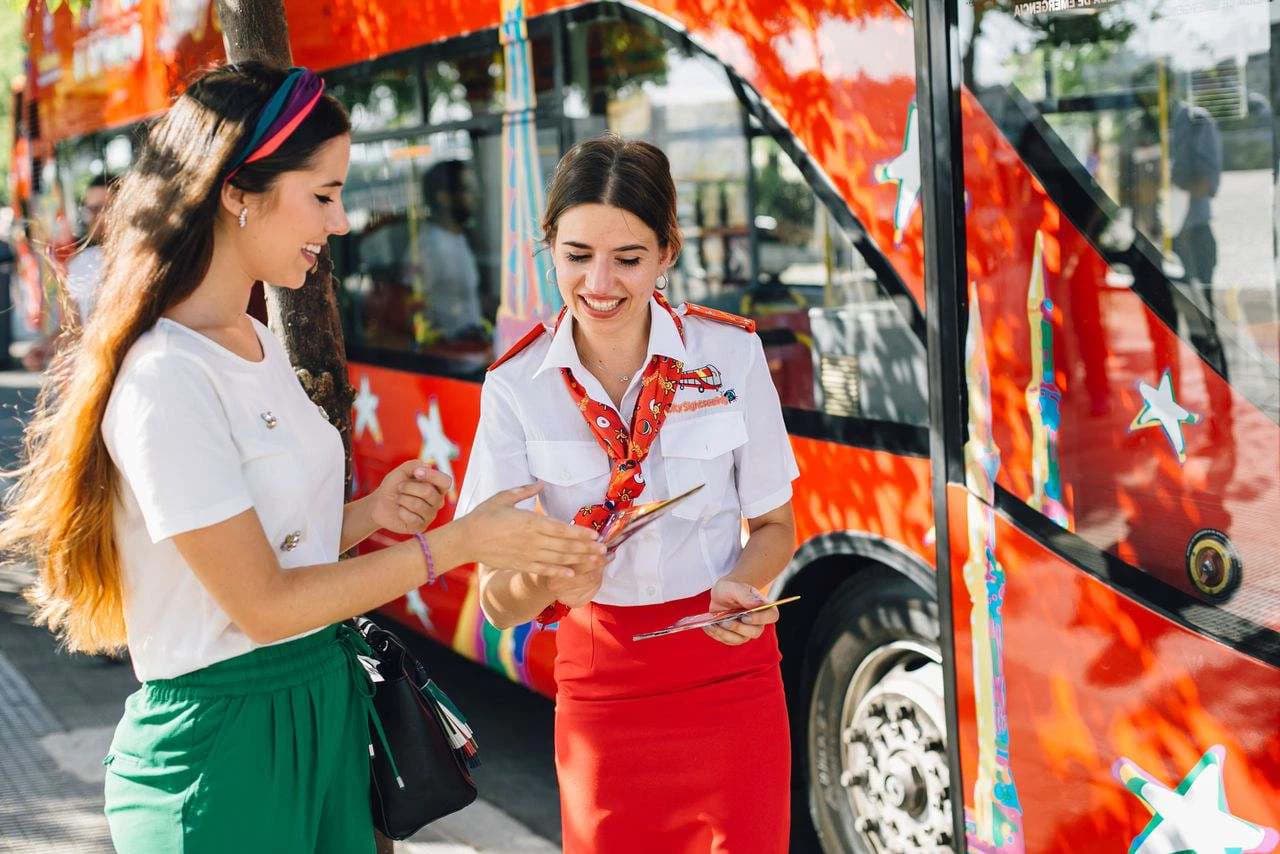 Two women smiling near a colorful red tour bus. One is in a uniform holding brochures, and the other is in casual attire