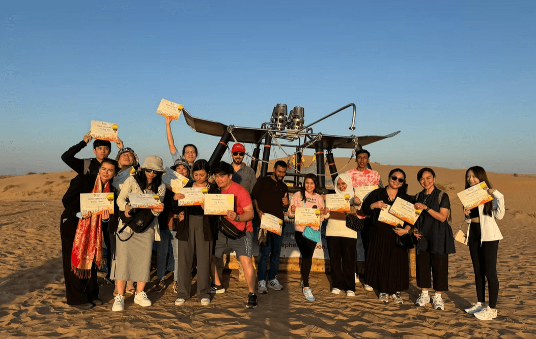 A group of people smiling and holding certificates stands together on a sandy desert landscape near a hot air balloon.