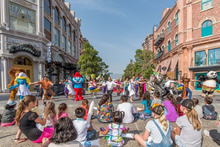 Street performance with colorful costumed characters, including mascots, entertaining an audience of children sitting on the ground.