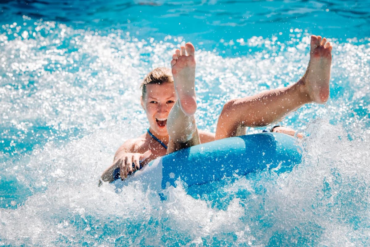A person joyfully rides a blue inner tube down a waterslide, splashing water everywhere.