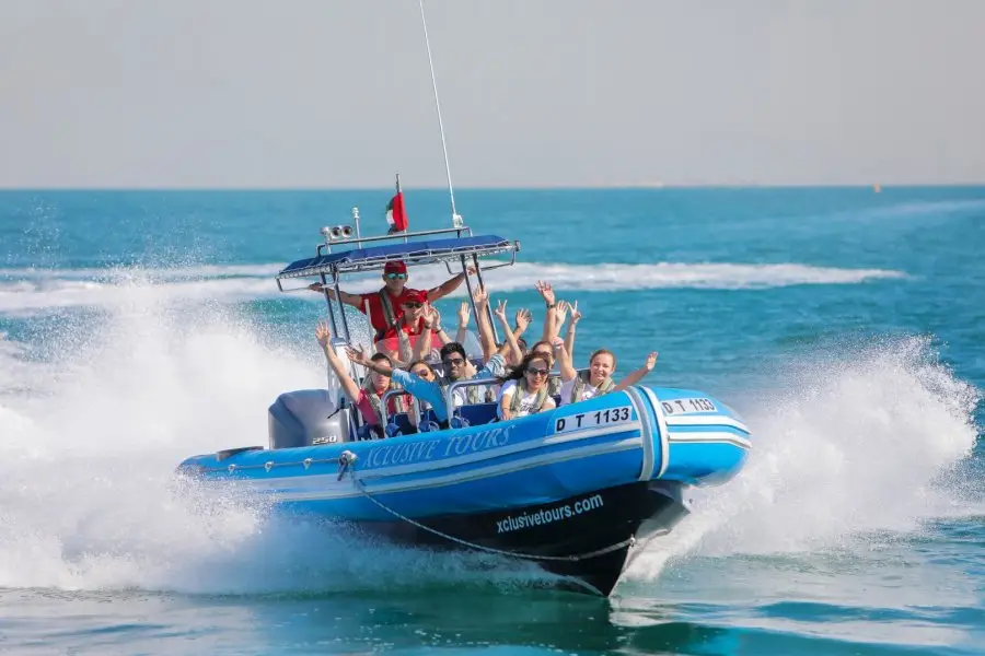 A blue speedboat cuts through bright blue water, scattering white spray. Passengers smiling and raising hands