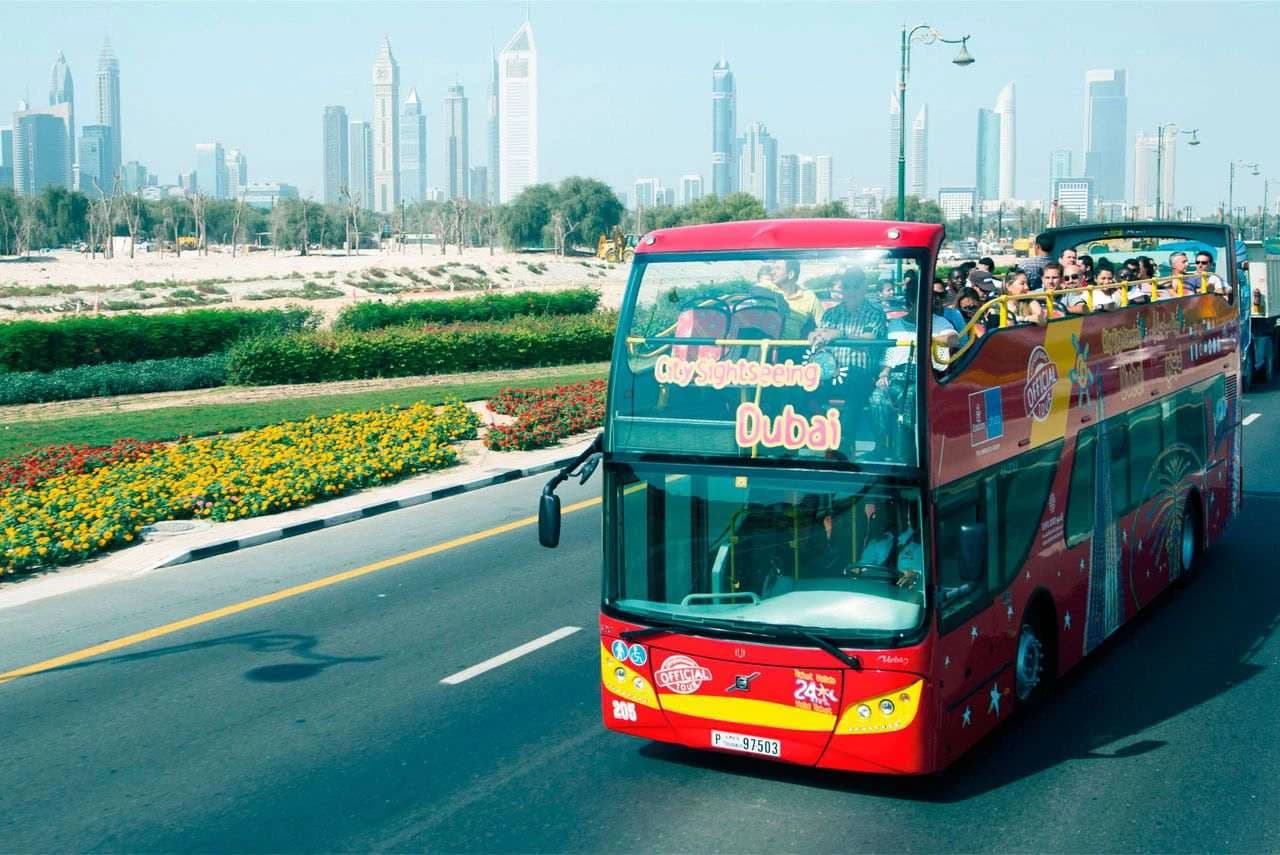 A red double-decker sightseeing bus with tourists on the open top drives along a road in Dubai