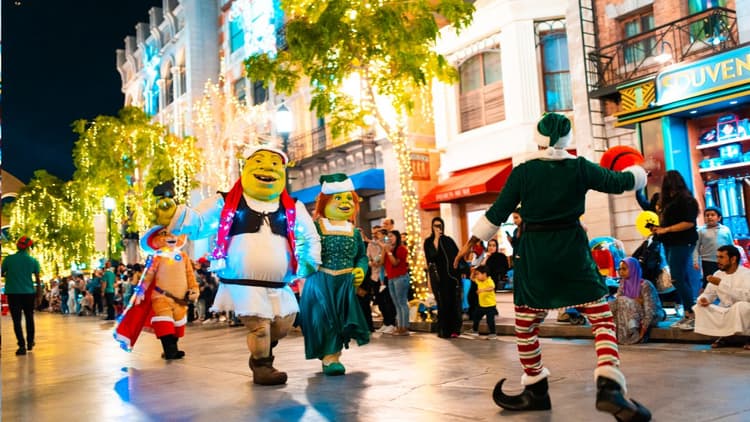 Street parade at night features vibrant Shrek and Fiona costumes, colorful lights on trees.