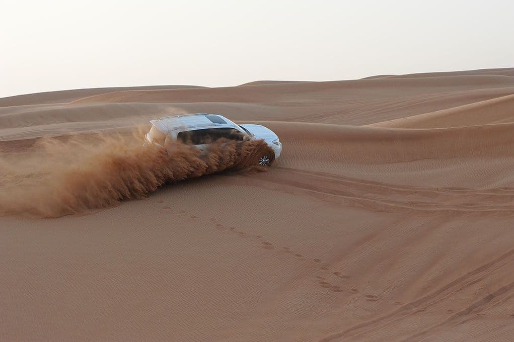 A white SUV navigates a sandy desert, kicking up a dramatic spray of sand. 