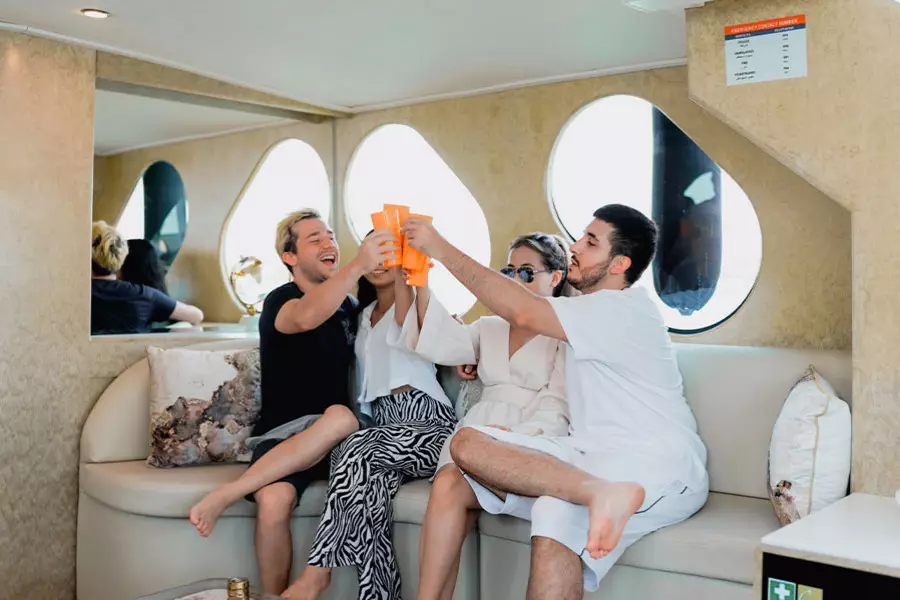 Four people sit on a yacht's sofa, smiling and raising orange drinks in a cheerful toast. 