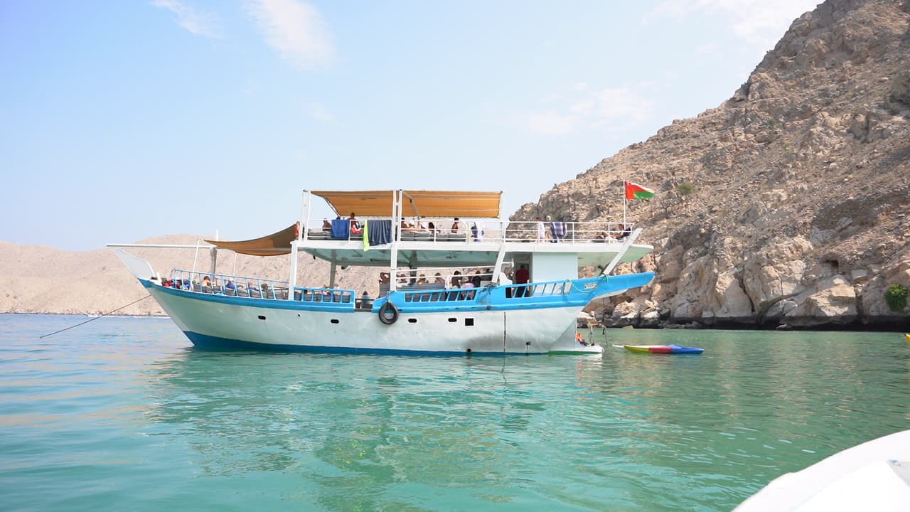 A large white and blue boat with orange canopies sits on calm green water, near rocky cliffs under a clear sky. 