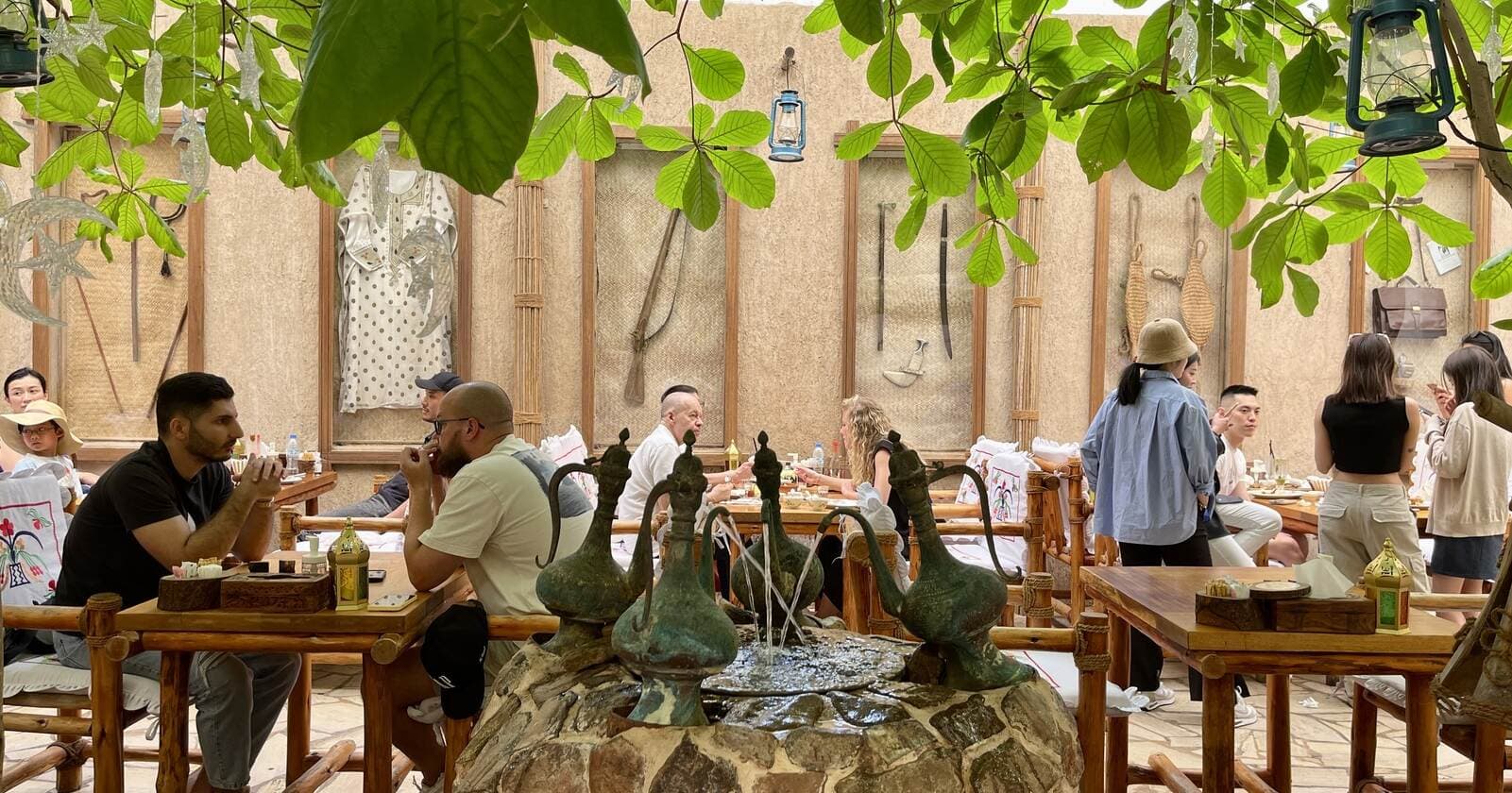 A cozy outdoor café with leafy trees overhead, wooden benches, and a central stone fountain. 