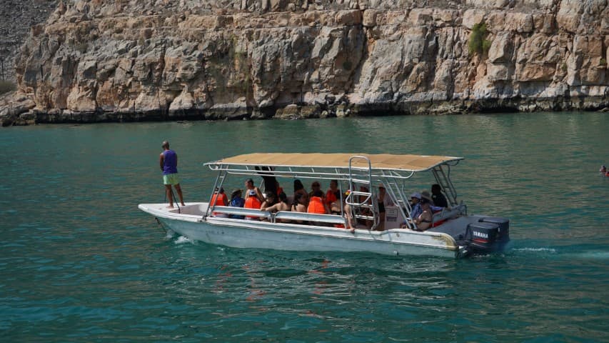 A boat with a beige canopy carries passengers in orange life vests on turquoise water.