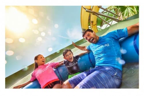 A family of four joyfully slides down a water slide at Dolphin Kayak Dubai, splashing into the water below.