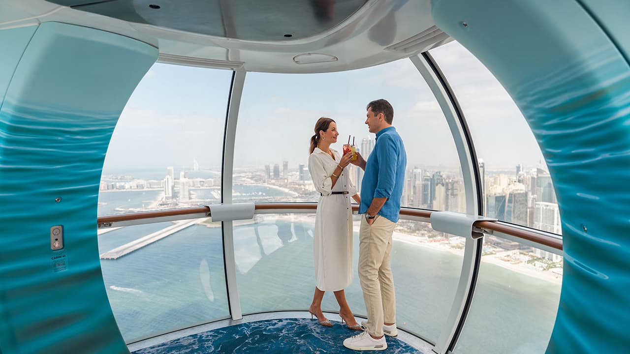 Couple enjoying drinks inside an Ain Dubai observation cabin with panoramic views of Dubai coastline and skyline.
